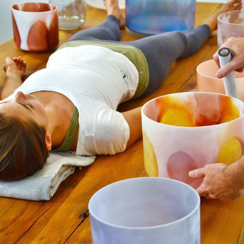 A lady is lying down receiving crystal bow therapy. Crystal bowls are surrounding her and a therapist is playing them.